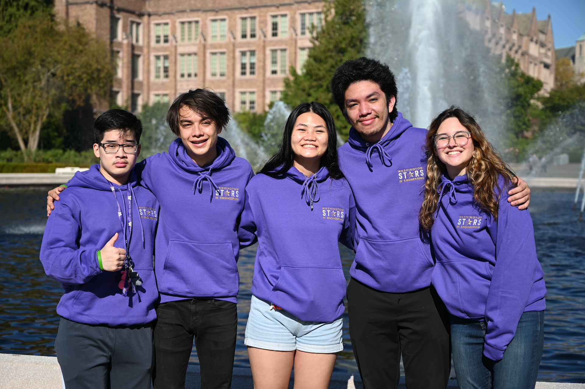 A group of students wearing purple UW hoodies and smiling at the camera with a water fountain and building in the background