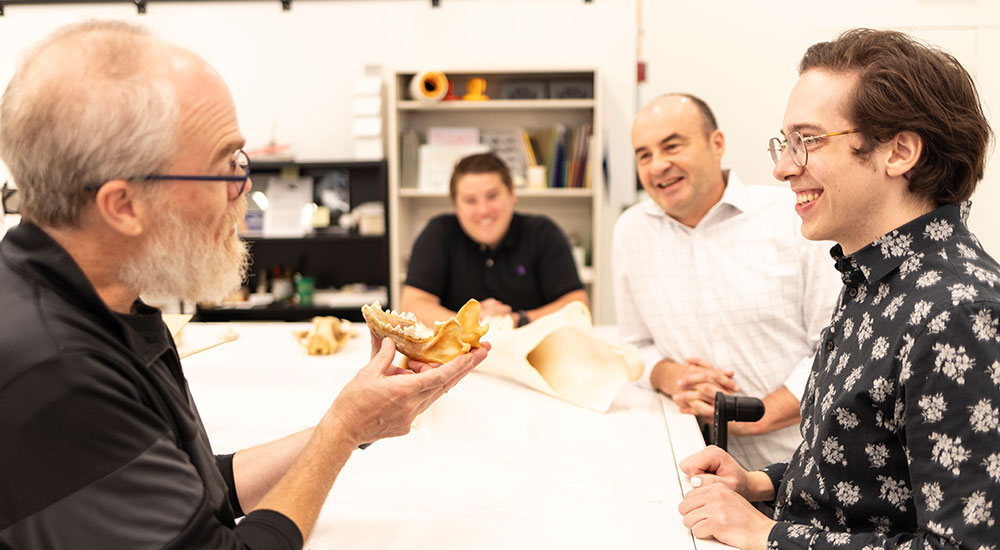 One person holds an African wild dog skull and is showing it to three other people sitting around table.