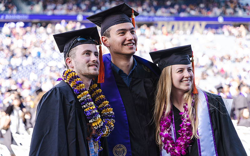 Three students wearing cap and gowns smiling for picture during commencement in Husky stadium