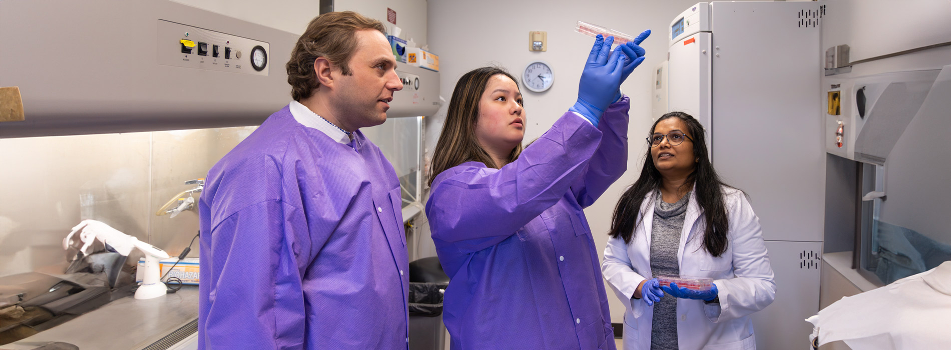 Three people in a laboratory, two in purple lab coats examining a petri dish, and one in a white lab coat observing.