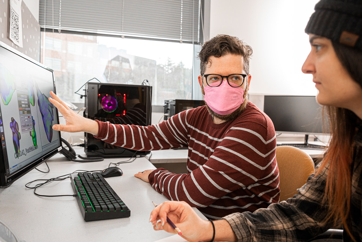 Two individuals working at a computer desk, one pointing at a screen displaying a 3D model.