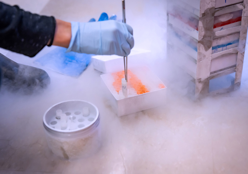 Laboratory scene with a gloved hand handling a sample from a box of vials amidst cold vapor.