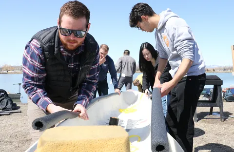 Two students stand on either side of a canoe and fasten foam insulation to the sides