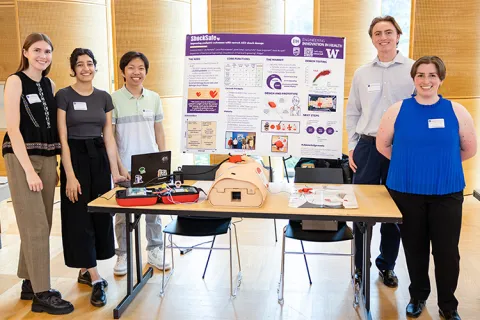 Five ShockSafe team members stand next to their poster and their prototype of their device