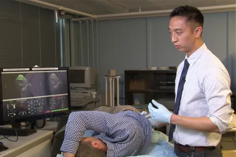 A man lies down in a hospital bed while another man holds a handheld device to his back while seeing ultrasound imaging on a computer screen.