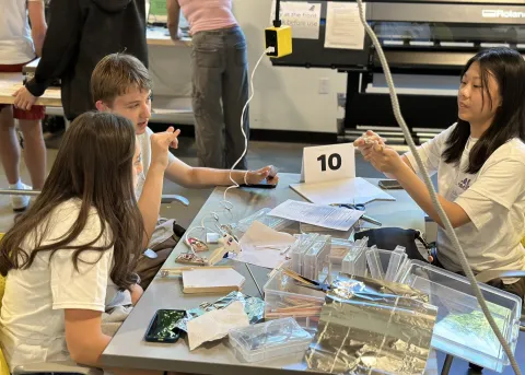 Engineering Academy students discuss a group project; one student points to another student holding a clothespin