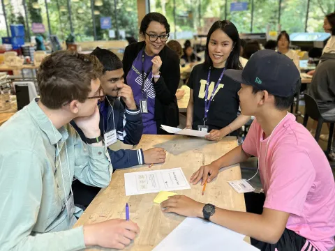 An engineering professor talks to three Engineering Academy students sitting at a workbench