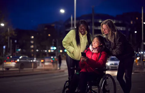 A person sitting in a wheelchair looking at a phone while two people are looking over her shoulder at the phone