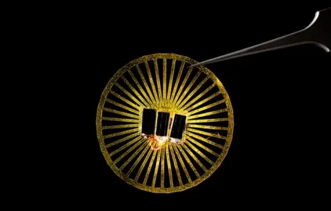 Tweezers holding a circular device with solar panels on it. It's illuminated against a black background.