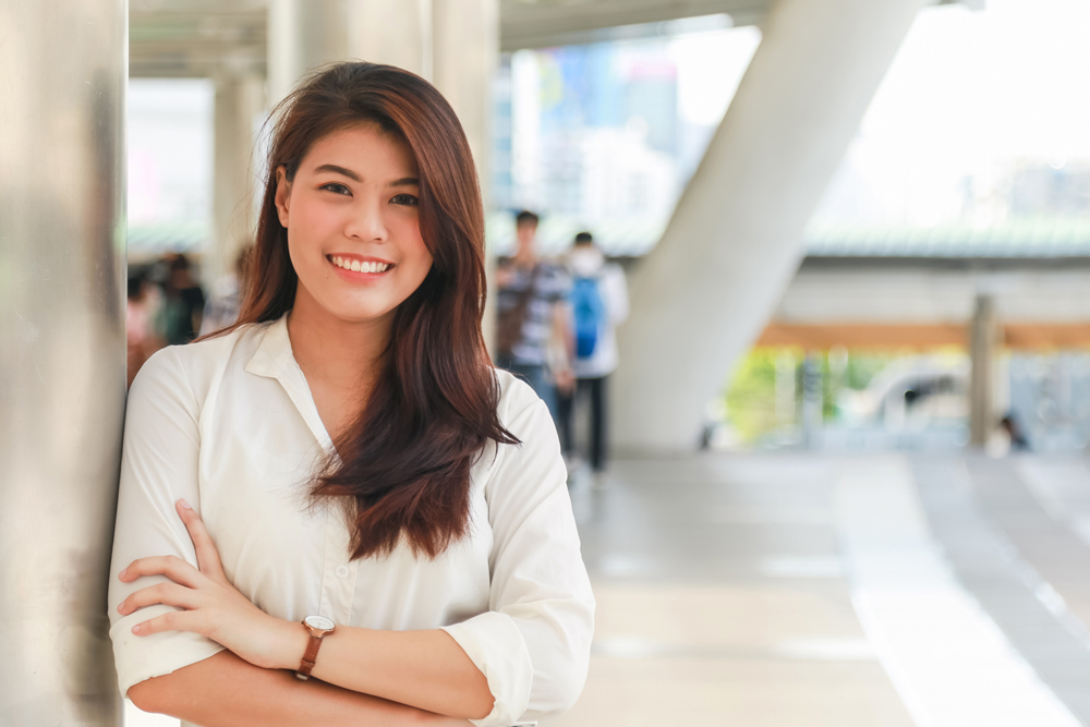 Smiling young woman in a white blouse leaning against a silver pole with arms crossed.