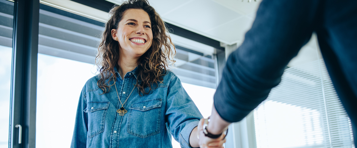 Woman in a blue denim shirt smiling while shaking hands with another person.