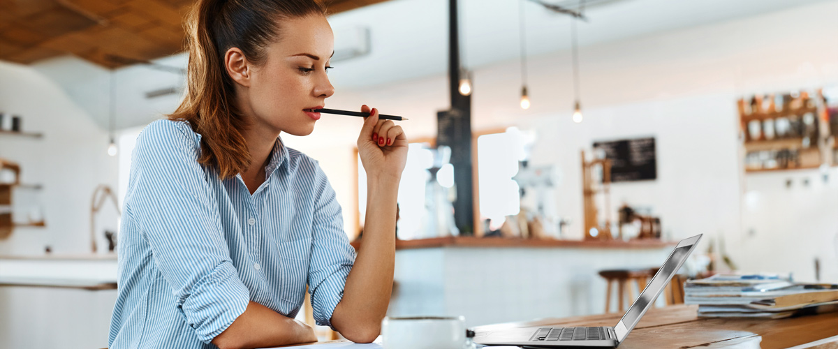 Woman in a café contemplating while looking at a laptop, holding a pen to her lips.