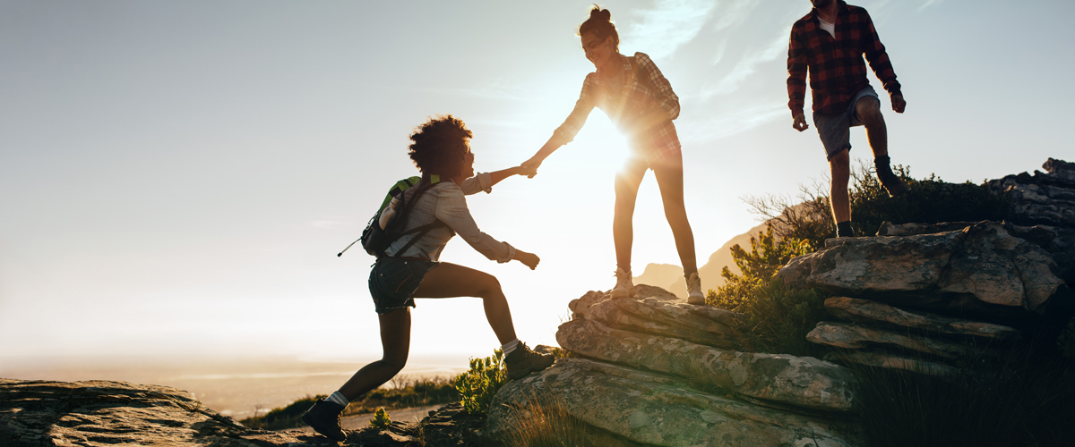 Three people hiking, one helping another climb a rocky outcrop during sunset.