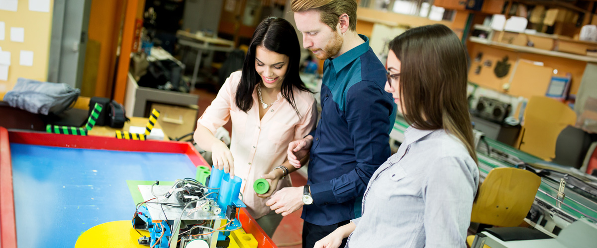 Three people working together on a robotic project at a workbench in a workshop.