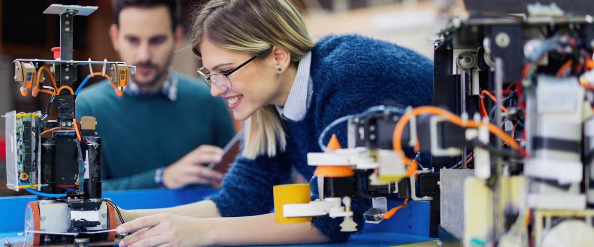 Woman working on a mechanical device with wires and circuit boards. Another person is in the background.