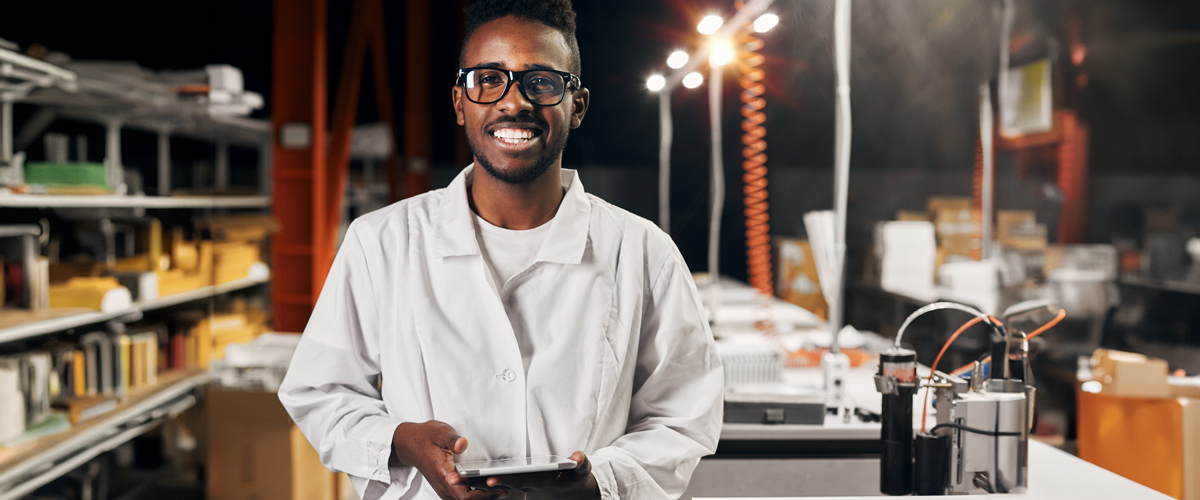  Man in a white lab coat and glasses holds a tablet while standing in a well-lit laboratory or workshop.