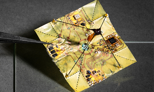 Close-up of a square electronic component held by a tweezer, showing intricate circuitry on a flexible material.