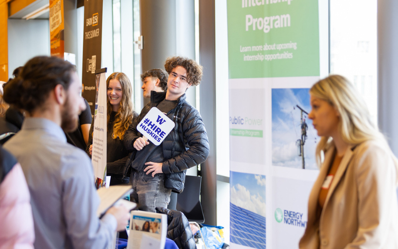 A busy career fair. In the background, a person is holding up a sign that reads #HireHuskies