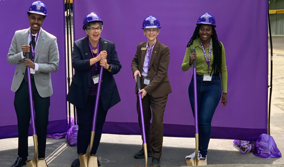 A ceremonial groundbreaking with four people holding shovels and a dog lying in front.