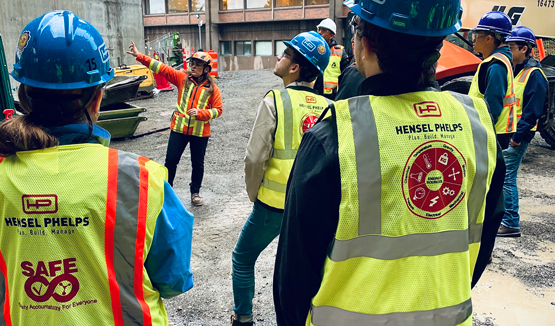 Construction workers in hard hats and safety vests at a worksite meeting, with one person gesturing.