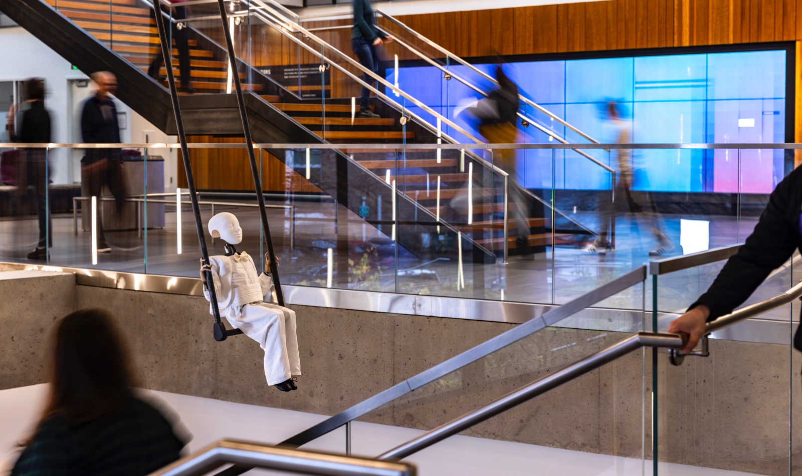 Not Afraid of Falling - A mannequin in white clothing seated on a swing over a stairway indoors.