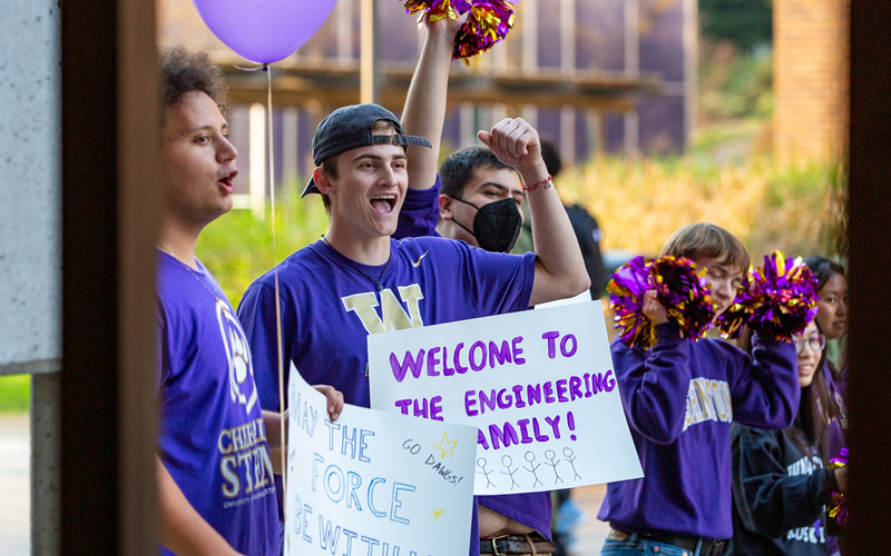 Students wearing purple and gold UW gear cheering on holding posters that say 'Welcome to the engineering family'