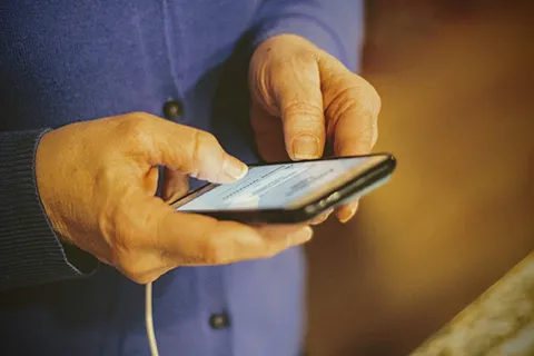 An older person's hands hold a smart phone. The person is wearing a blue sweater.