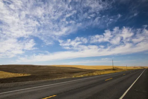 Two-lane road with yellow lines flanked by plowed and grassy fields under a blue sky with scattered clouds.