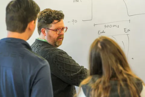 Person pointing at writing on whiteboard while two individuals watch