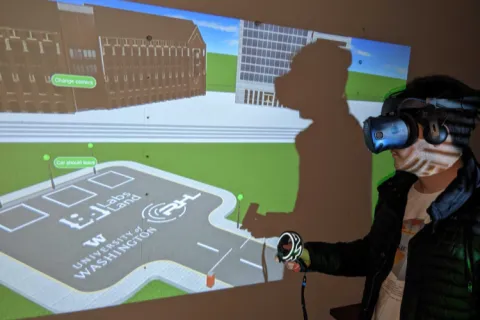 A person with a virtual reality helmet on stands in front of a screen that showcases a virtual world with two buildings and a parking lot