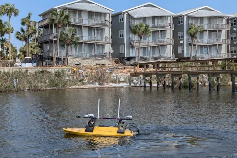 A yellow aquatic drone on a body of water with residential buildings and palm trees in the background