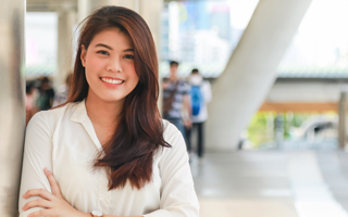 A young woman with long brown hair, wearing a white shirt, smiling and standing on a pedestrian walkway.